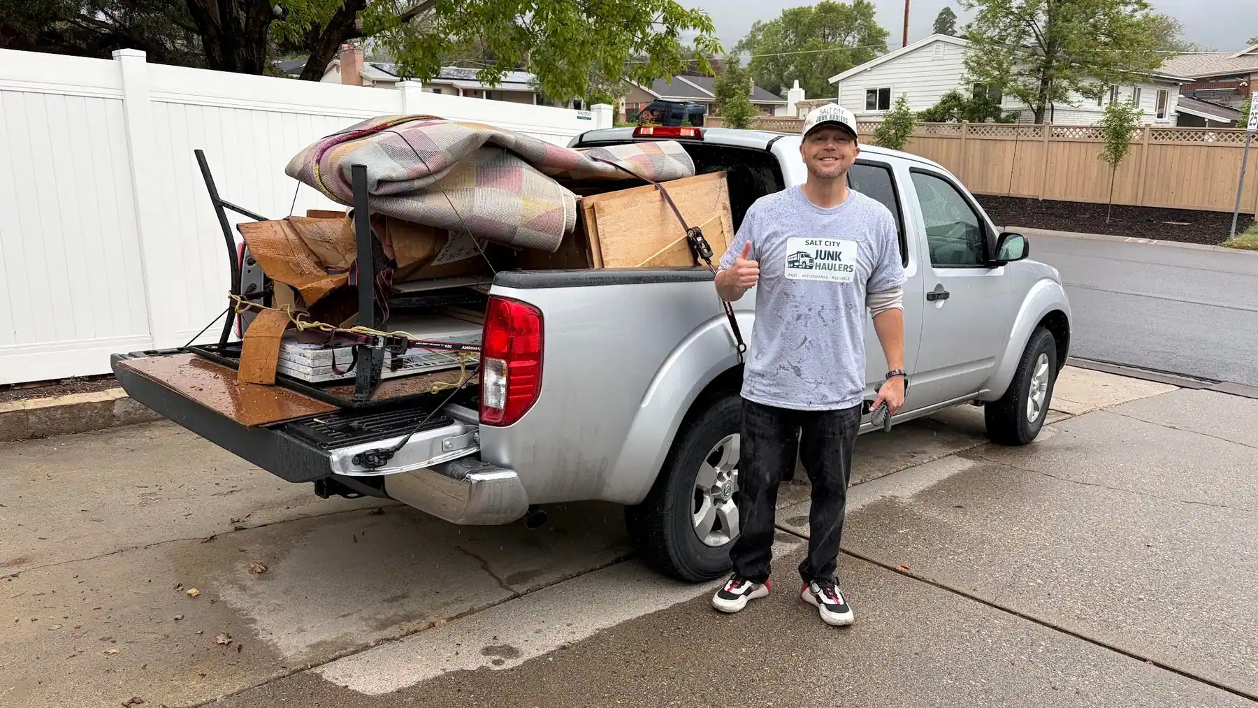 Jeremy Saxey giving a thumbs up next to his silver pickup loaded with a mattress, a bed frame and old furniture on a Salt Lake City driveway